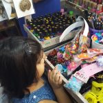 A young girl wearing a blue dress is looking at various colorful accessories and toys displayed on a market stall. The table is crowded with hair clips, nail polish bottles, decorative items, and other small trinkets.