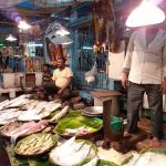 A bustling fish market scene with fish displayed on banana leaves. Two vendors sit behind the counter while a standing vendor handles a cleaver. Various types of fish are laid out for sale, with colorful decorations and lights hanging from the ceiling in the background.