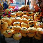 A stall displays a variety of yellow emoji cushions with different facial expressions. The collection includes smiles, winks, kisses, and laughter emojis. The bright and colorful setup also features plush toys in the background, creating a cheerful atmosphere.