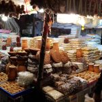 A bustling market stall is filled with a variety of items, including grains, spices, snacks, and earthenware. The stall is brightly lit and packed with a diverse assortment of products, with stacks of bags and containers displayed prominently.