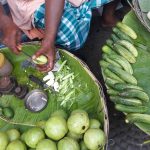 A vendor seated with baskets of fresh vegetables. They are cutting a cucumber on a banana leaf with a knife. Surrounding them are guavas, cucumbers, and other produce. The baskets are lined with banana leaves. The vendor is wearing a checkered lungi.