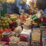 A vibrant market stall displaying a variety of fresh fruits, packaged foods, and spices. Bananas, custard apples, and other fruits are prominent alongside stacks of boxed and bagged items. The stall is bustling, well-lit, and densely packed with goods.