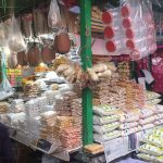 A bustling market stall with various packaged goods, including cereals, lentils, and snacks, displayed in heaps. Transparent plastic bags with items hang from the green framework. Shoppers browse the vibrant stall, which is surrounded by colorful fabrics and bags.