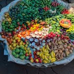 A large, round basket is filled with an array of colorful vegetables, fruits, and clay pots. The produce includes tomatoes, bananas, green beans, potatoes, and more, arranged in a visually appealing manner on a textured floor. There's a colorful plate on top.