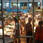 A bustling fish market scene with vendors and customers moving around. Various types of fish and seafood are displayed on tables. The market is crowded, with people conversing and inspecting the goods. Bright lights illuminate the lively atmosphere.