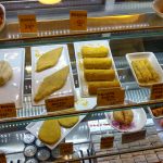 Displays various fried items in a bakery. Labeled pastries, sandwiches, various breaded items, and samosas are arranged on white plates in a glass case. Small signs with prices are placed next to the food items.