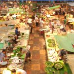 A busy market with several stalls selling fresh fish and seafood. Vendors are seen engaging with customers amidst an array of colorful fish laid out on tables. Green tarps cover some stalls, and the market is bustling with activity and vibrant colors.