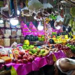 A vibrant fruit and spice market stall is bursting with a colorful array of fresh produce, including melons, apples, bananas, and mangoes. Various nuts, spices, and dried fruits are neatly displayed in jars and plastic bags, lit by white lamps. A vendor is partially seen handling goods.