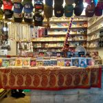 A small shop selling a variety of goods, including hats displayed on the upper racks, DVDs with covers featuring various films on the counter, colorful strings, and other trinkets hanging from hooks. The shopkeeper, partially visible, is seated behind the counter.