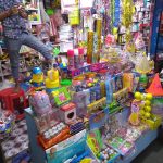 A colorful shop stall filled with a variety of toys and celebration items, including party hats, decorative items, balloons, and stationery. A person, partially visible, standing on a red stool and arranging items on the shelves. The shop's interior is densely packed.