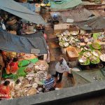 A bustling fish market scene from above shows various vendors with tables full of different types of fish and seafood. People are seen examining the produce, and some are haggling. The market area is covered with makeshift tarps, and the floor is wet.