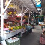 A narrow market street lined with various fruit and vegetable stalls. Fresh produce including bananas, melons, apples, and other colorful fruits are displayed. Customer and vendor interactions can be seen in the background under a covering.