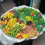 A large round tray, placed on the ground, is filled with a colorful assortment of small fruits and vegetables at a market stall. The produce includes various peppers, citrus fruits, eggplants, okra, and other exotic items, arranged in a visually appealing manner.